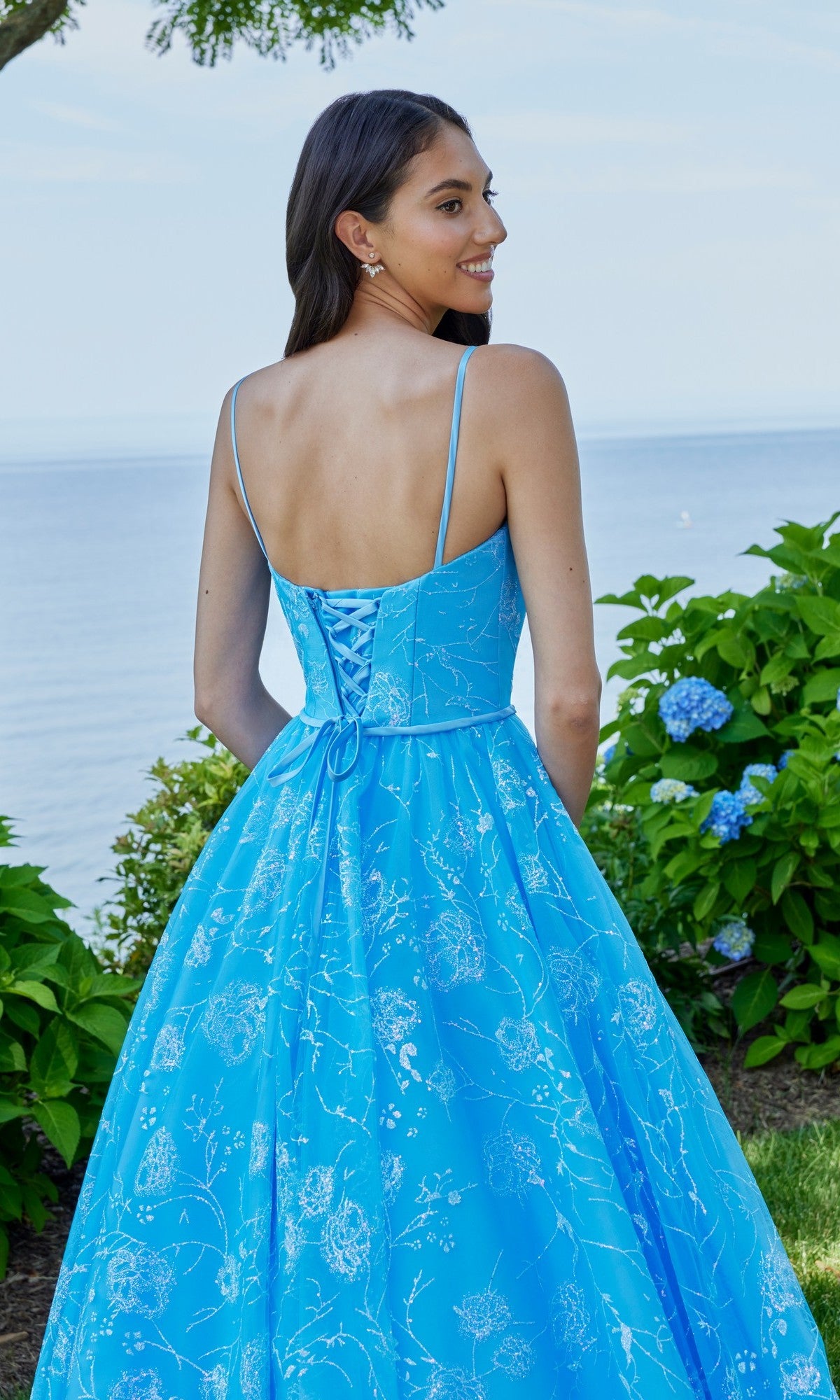 A woman stands outdoors near green bushes and blue flowers, wearing the Morilee 49153 long prom dress in blue with a sleeveless A-line design and lace-up back, with water visible in the background.
